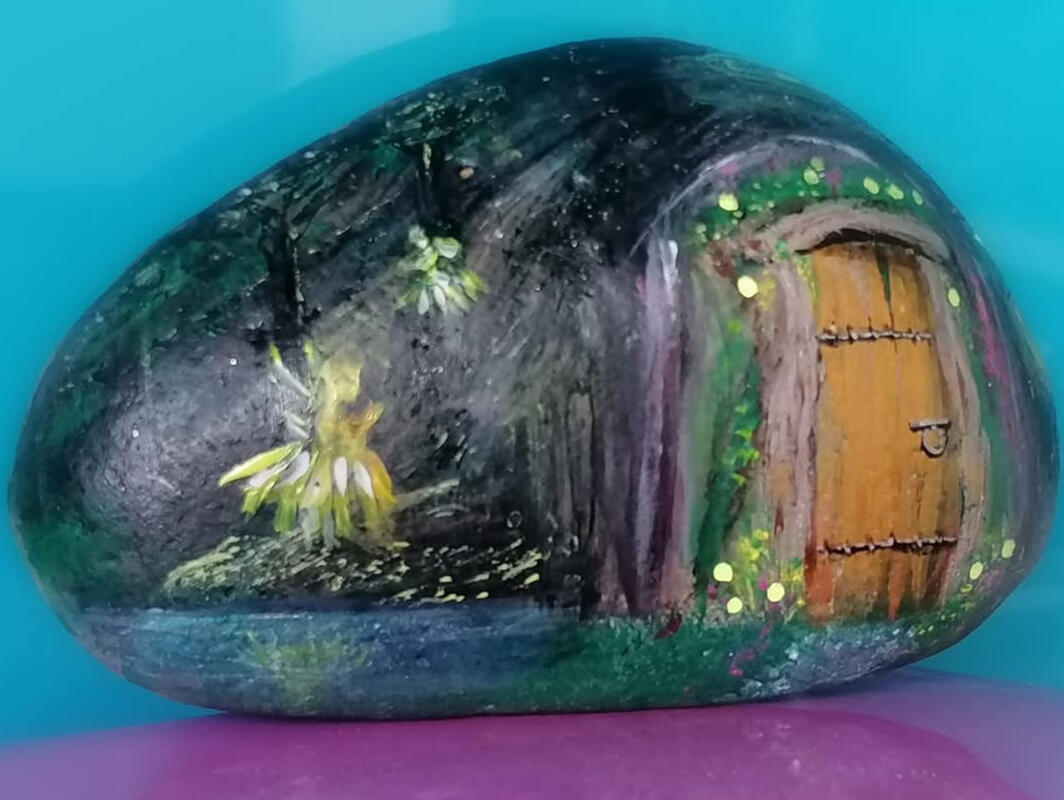 A hand painted stone with an ethereal fairy in a forest by an ornate wooden door surrounded by flowers. By Sue's Stones and Fine Art